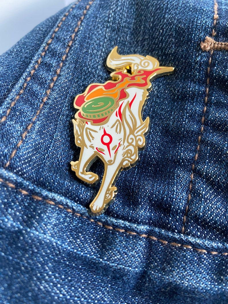 May include: A gold-colored enamel pin featuring a white fox with red markings. The fox is standing on its hind legs with its tail curled up. The fox is wearing a green and red hat with a gold trim. The pin is on a blue denim background.