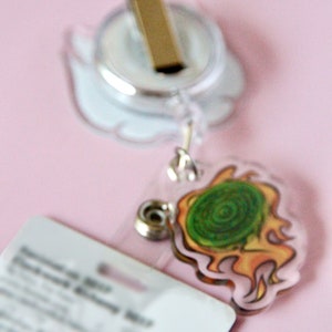 May include: A clear plastic badge holder with a retractable reel and a gold clip. The holder has a green and orange cartoon design of a swirling vortex.