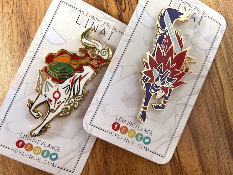 May include: Two enamel pins featuring stylized wolf designs. The first pin is white with red and gold accents and depicts a wolf with a red and green flame behind it. The second pin is purple with red and gold accents and depicts a wolf with a sword behind it. Both pins have the text "LINAIREYLANCE" and social media icons on the back.