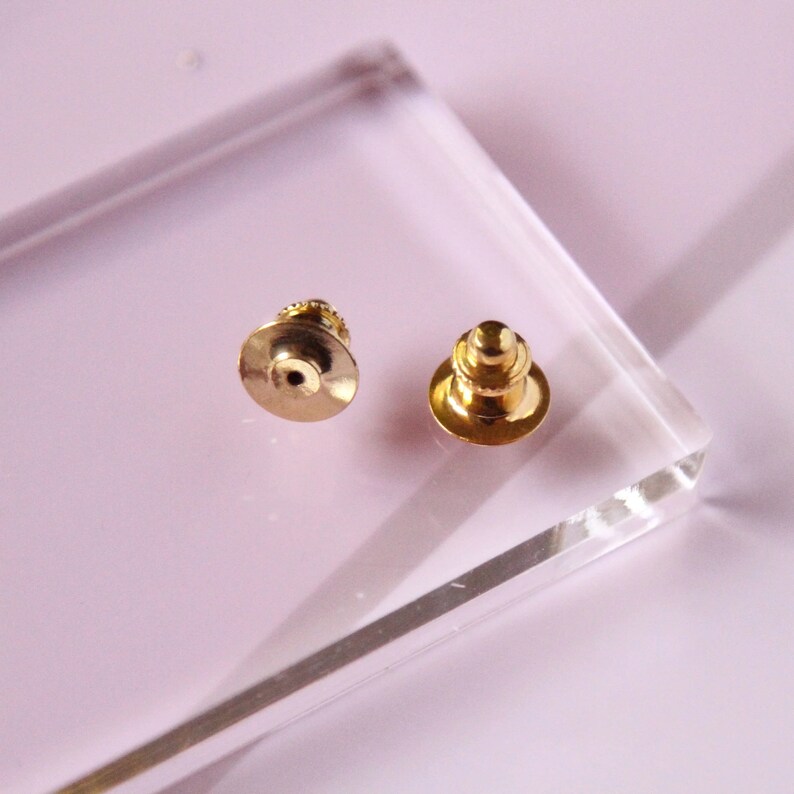 Op de afbeelding: Twee gouden metalen veiligheidsspeldruggen. De veiligheidsspeldruggen hebben een ronde kop en een puntig uiteinde.