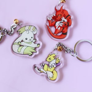 May include: Three acrylic keychains featuring cartoon characters from the Digimon anime series. The keychains are on a pink background. The characters are a yellow and white fox-like creature, a red and black dragon-like creature, and a white and green dog-like creature.