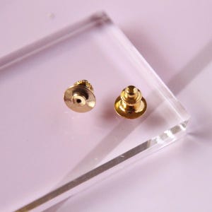 May include: Two gold-toned metal pin backs with a round base and a pointed top. The pin backs are on a clear acrylic surface.
