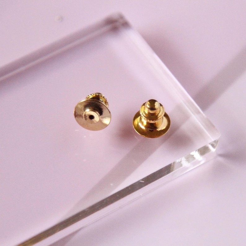 May include: Two gold-toned metal pin backs with a round base and a pointed top. The pin backs are on a clear acrylic surface.