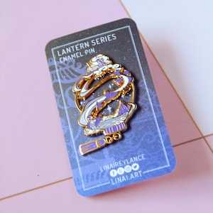 Peut inclure: Une épingle en émail dorée de la "Série Lanterne" avec un motif de lanterne stylisée avec des accents blancs, violets et bleus. L'épingle est sur une carte bleu foncé avec le texte "LANTERN SERIES ENAMEL PIN".