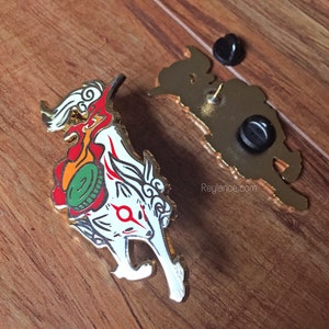May include: A gold-colored enamel pin featuring a white fox with red and orange flames, a green and gold coin, and a red circle on its forehead. The pin has a gold-colored back with a safety pin closure and the text "Reylance.com".