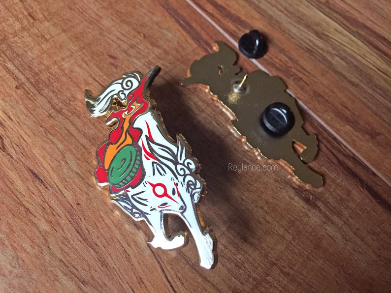 May include: A gold-colored enamel pin featuring a white fox with red and orange flames, a green and gold coin, and a red circle on its forehead. The pin has a gold-colored back with a safety pin closure and the text "Reylance.com".