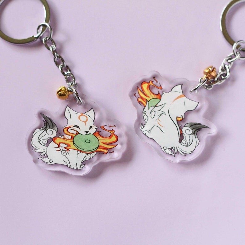 May include: Two white fox keychains with orange flames and a green circle. One fox is holding the circle and the other is sitting with its tail curled around its legs.