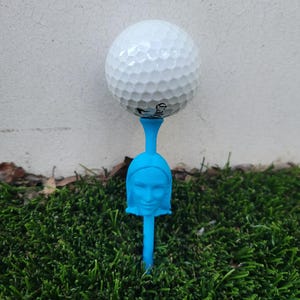 Puede incluir: Una pelota de golf blanca está equilibrada sobre un tee de golf azul. El tee presenta un diseño de rostro esculpido. El tee está colocado sobre césped artificial verde. El fondo es una pared clara.