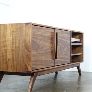 May include: A mid-century modern walnut wood entertainment center with two doors and open shelves. The cabinet has a rich brown color and features a unique design with angled legs.