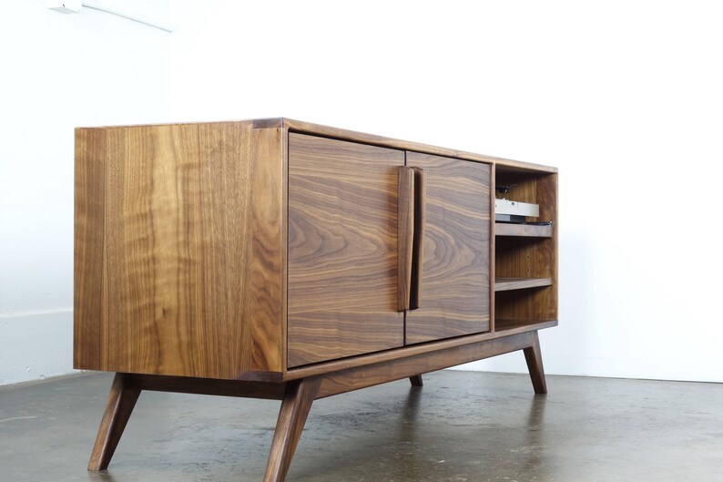 May include: A mid-century modern walnut wood entertainment center with two doors and open shelves. The cabinet has a rich brown color and features a unique design with angled legs.