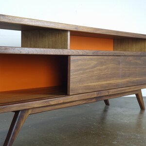 Puede incluir: Un centro de entretenimiento de estilo moderno de mediados de siglo en madera de nogal con una puerta corredera y estantes abiertos. Los estantes est&aacute;n pintados de naranja.