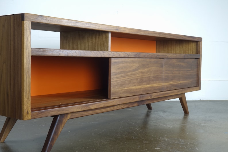 Puede incluir: Un centro de entretenimiento de estilo moderno de mediados de siglo en madera de nogal con una puerta corredera y estantes abiertos. Los estantes est&aacute;n pintados de naranja.