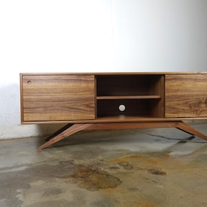 Puede incluir: Un centro de entretenimiento moderno de estilo mediados de siglo hecho de madera de nogal, con dos puertas y un estante. El centro de entretenimiento tiene patas inclinadas y un dise&ntilde;o minimalista.