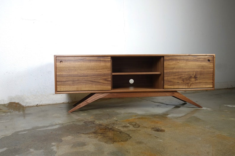 Puede incluir: Un centro de entretenimiento moderno de estilo mediados de siglo hecho de madera de nogal, con dos puertas y un estante. El centro de entretenimiento tiene patas inclinadas y un dise&ntilde;o minimalista.