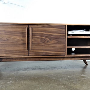 May include: A mid-century modern walnut wood entertainment center with two doors and open shelving. The entertainment center has a record player on the top shelf and a cutout for cords on the bottom shelf.