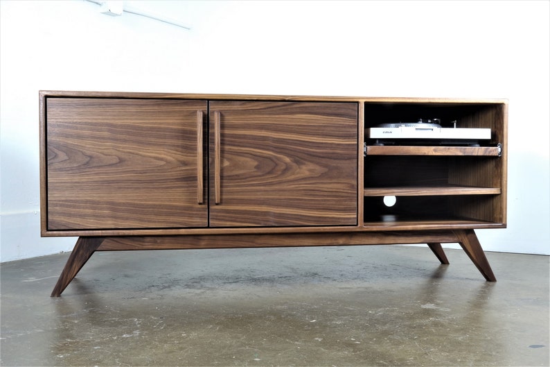 May include: A mid-century modern walnut wood entertainment center with two doors and open shelving. The entertainment center has a record player on the top shelf and a cutout for cords on the bottom shelf.