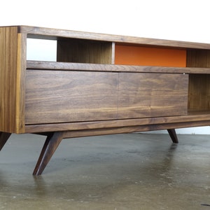 Puede incluir: Un centro de entretenimiento de estilo moderno de mediados de siglo en madera de nogal con una puerta corredera, estantes abiertos y patas inclinadas. El centro tiene un acento naranja brillante en la pared posterior.