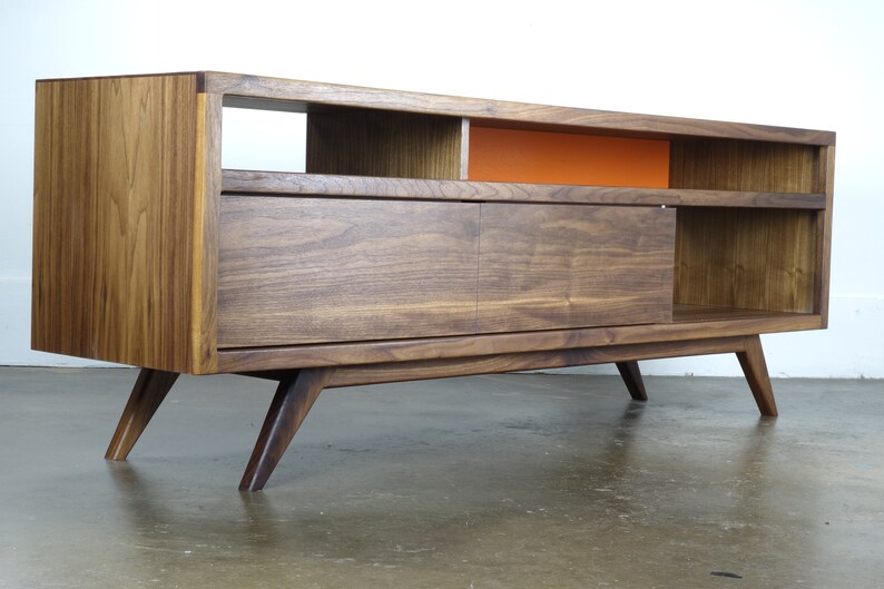 Puede incluir: Un centro de entretenimiento de estilo moderno de mediados de siglo en madera de nogal con una puerta corredera, estantes abiertos y patas inclinadas. El centro tiene un acento naranja brillante en la pared posterior.