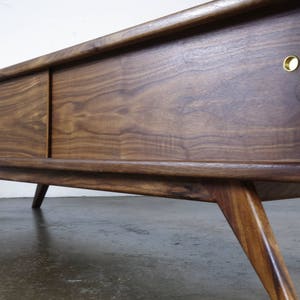 May include: A mid-century modern walnut wood cabinet with sliding doors and brass hardware. The cabinet has a rich brown color and a sleek design. It is sitting on a light gray floor.