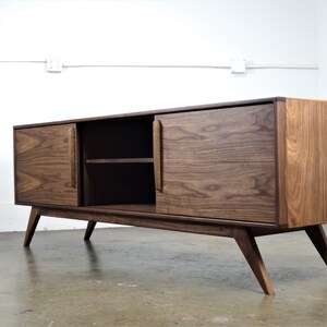 May include: A mid-century modern style walnut wood media console with two doors and a shelf. The console has a dark brown finish and features tapered legs.