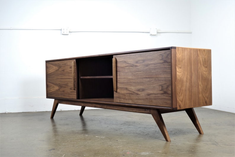 May include: A mid-century modern style walnut wood media console with two doors and a shelf. The console has a dark brown finish and features tapered legs.