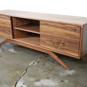 Puede incluir: Un centro de entretenimiento largo y rectangular de madera de nogal con dos puertas correderas y dos estantes. El centro de entretenimiento tiene patas inclinadas.