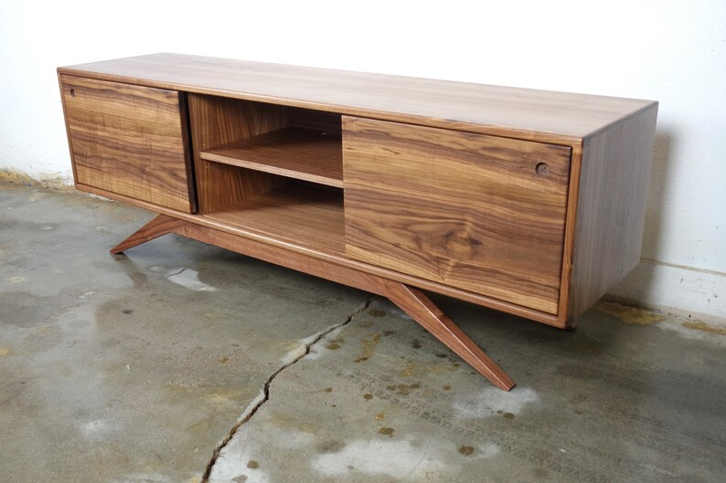 Puede incluir: Un centro de entretenimiento largo y rectangular de madera de nogal con dos puertas correderas y dos estantes. El centro de entretenimiento tiene patas inclinadas.