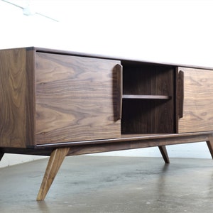 May include: A mid-century modern walnut wood media console with two sliding doors and a shelf. The console has a dark brown finish and sits on four angled legs.