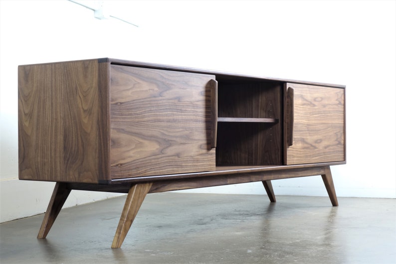May include: A mid-century modern walnut wood media console with two sliding doors and a shelf. The console has a dark brown finish and sits on four angled legs.