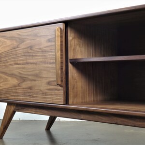 May include: A mid-century modern walnut wood entertainment center with a single door and open shelving. The cabinet has a unique angled leg design and a small hole in the bottom shelf for cord management.