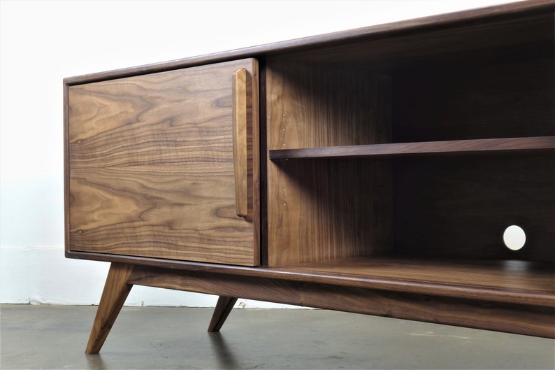 May include: A mid-century modern walnut wood entertainment center with a single door and open shelving. The cabinet has a unique angled leg design and a small hole in the bottom shelf for cord management.