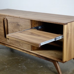 May include: A mid-century modern style walnut wood entertainment console with two doors and a pull-out shelf. The console has a light brown finish and sits on four angled legs.