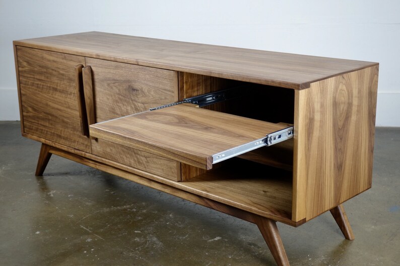 May include: A mid-century modern style walnut wood entertainment console with two doors and a pull-out shelf. The console has a light brown finish and sits on four angled legs.
