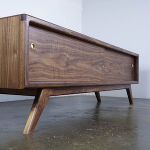 May include: A mid-century modern walnut wood entertainment center with a sliding door and brass hardware. The cabinet has a dark wood finish and sits on four angled legs.