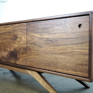 Puede incluir: Un mueble de TV moderno de madera de nogal de estilo mediados de siglo con dos puertas correderas y un estante. El mueble tiene patas inclinadas y un acabado marr&oacute;n oscuro.