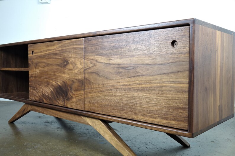 Puede incluir: Un mueble de TV moderno de madera de nogal de estilo mediados de siglo con dos puertas correderas y un estante. El mueble tiene patas inclinadas y un acabado marr&oacute;n oscuro.