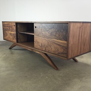 Puede incluir: Un centro de entretenimiento de madera de nogal de estilo mediados de siglo moderno con puertas correderas y un estante. El gabinete tiene un acabado marr&oacute;n oscuro y patas anguladas.