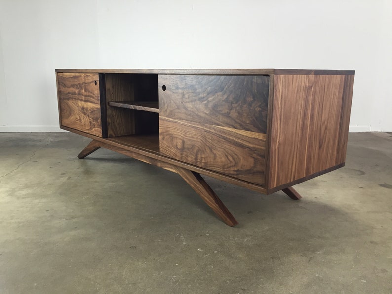 Puede incluir: Un centro de entretenimiento de madera de nogal de estilo mediados de siglo moderno con puertas correderas y un estante. El gabinete tiene un acabado marr&oacute;n oscuro y patas anguladas.