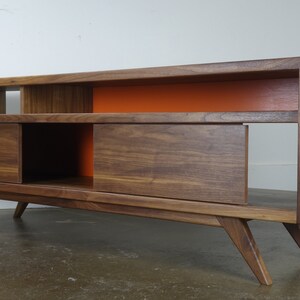 Puede incluir: Un centro de entretenimiento de estilo moderno de mediados de siglo con un acabado en madera de nogal. La unidad cuenta con dos puertas correderas, estantes abiertos y un acento naranja brillante en la pared posterior. El centro de entretenimiento tiene cuatro patas que se inclinan hacia afuera.