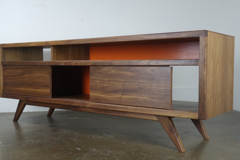 Puede incluir: Un centro de entretenimiento de estilo moderno de mediados de siglo con un acabado en madera de nogal. La unidad cuenta con dos puertas correderas, estantes abiertos y un acento naranja brillante en la pared posterior. El centro de entretenimiento tiene cuatro patas que se inclinan hacia afuera.
