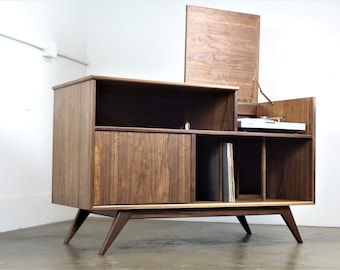 May include: A mid-century modern walnut record player console with a hinged lid, a record storage shelf, and a cabinet with a sliding door. The console has four legs and is made of solid wood.