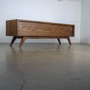 May include: A mid-century modern walnut wood media console with two doors and brass hardware. The console has angled legs and a sleek, minimalist design.