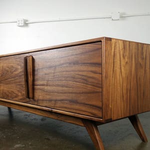 May include: A mid-century modern walnut wood entertainment console with two sliding doors and a shelf. The console has a rich brown color and a sleek design with angled legs.
