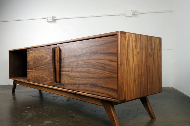 May include: A mid-century modern walnut wood entertainment console with two sliding doors and a shelf. The console has a rich brown color and a sleek design with angled legs.