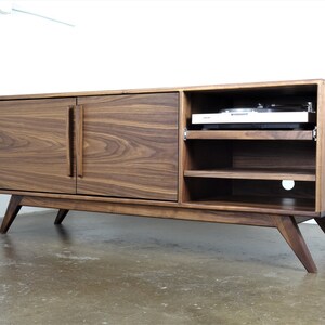 May include: A mid-century modern walnut wood entertainment center with two doors and open shelving. The unit has a record player on the top shelf and a cut-out for cords on the bottom shelf.