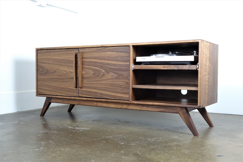 May include: A mid-century modern walnut wood entertainment center with two doors and open shelving. The unit has a record player on the top shelf and a cut-out for cords on the bottom shelf.