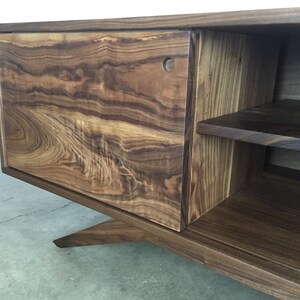 Puede incluir: Un centro de entretenimiento de madera marr&oacute;n oscuro con una puerta corredera y estantes abiertos. El centro de entretenimiento tiene una base angulada &uacute;nica.