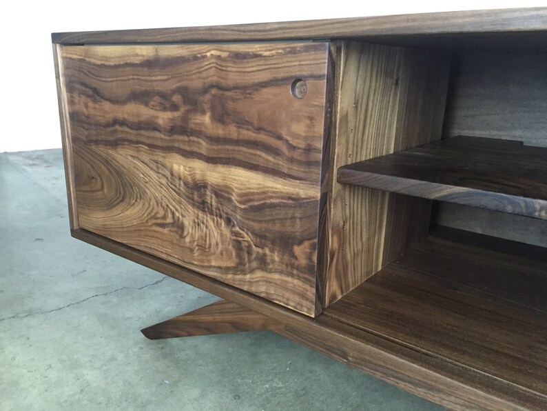 Puede incluir: Un centro de entretenimiento de madera marr&oacute;n oscuro con una puerta corredera y estantes abiertos. El centro de entretenimiento tiene una base angulada &uacute;nica.