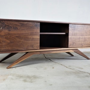 Puede incluir: Una consola de medios de madera de nogal de estilo moderno de mediados de siglo con puertas correderas y un estante. La consola tiene un acabado marr&oacute;n claro y se asienta sobre patas inclinadas.