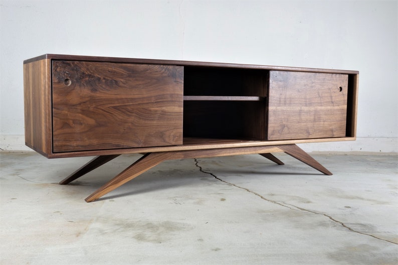 Puede incluir: Una consola de medios de madera de nogal de estilo moderno de mediados de siglo con puertas correderas y un estante. La consola tiene un acabado marr&oacute;n claro y se asienta sobre patas inclinadas.